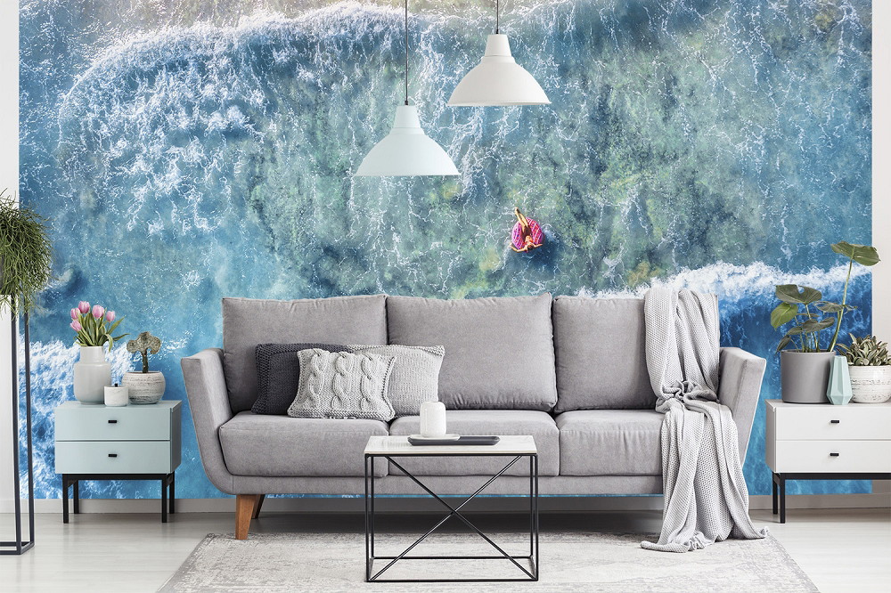 Fototapete - Wohnzimmer mit grauem Sofa und zwei hängenden Lampen, im Hintergrund eine Fototapete mit einer Luftaufnahme des Meeres und Wellen.