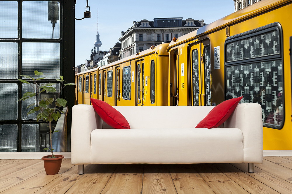 Fototapeta - Biała sofa z czerwonymi poduszkami przed dużym obrazem żółtego berlińskiego pociągu metra na fototapecie. Wieża telewizyjna i architektura Berlina w tle.