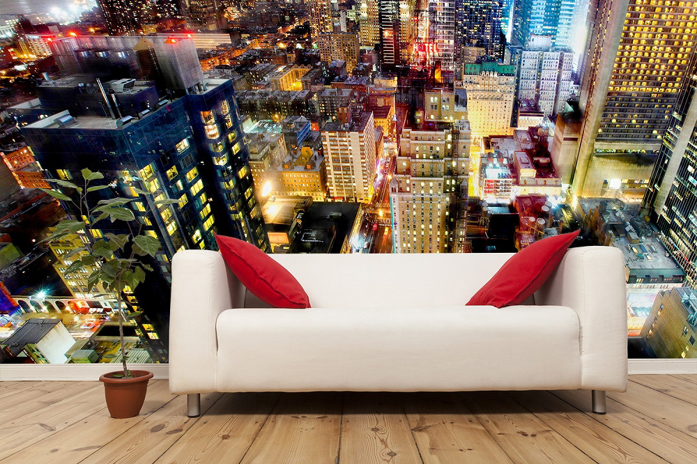 Fototapete - Abendlicher Stadtblick über eine beleuchtete urbane Skyline mit modernen Hochhäusern, hinter einem gemütlichen Wohnzimmer mit Sofa und Pflanzen