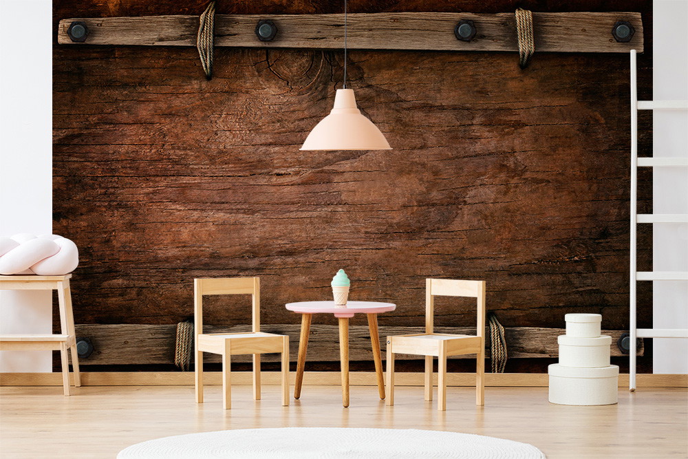 Fotomurale - Una parete rustica in legno fa da sfondo a un tavolino rotondo, due sedie per bambini ed elementi decorativi in una stanza luminosa che crea un'atmosfera invitante
