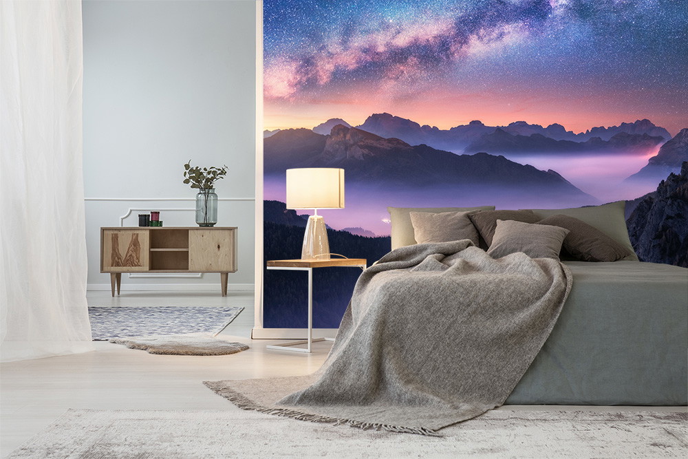 Fotobehang - Slaapkamer met bed en nachtkastje tegen een indrukwekkende sterrenhemel en bergpanorama