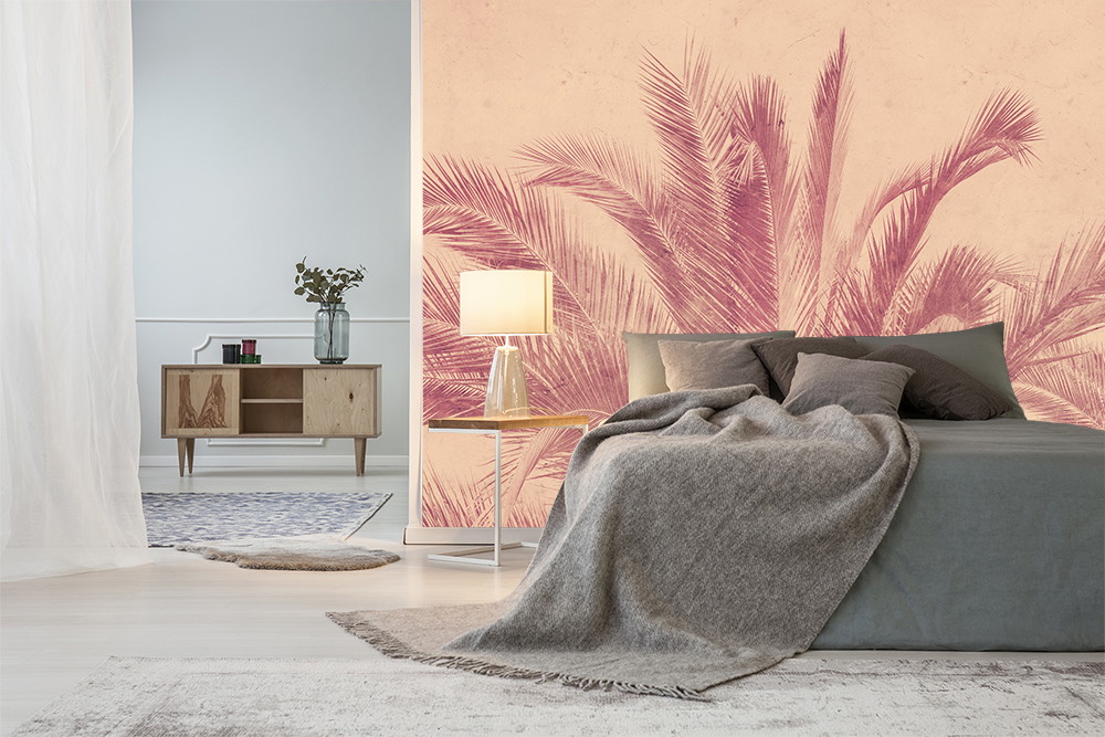 Fototapete - Schlafzimmer mit einer großen Palmen-Tapete in Rosatönen, einem grauen Bett mit Decke, Nachttischlampe und minimalistischem Möbelstück im Hintergrund