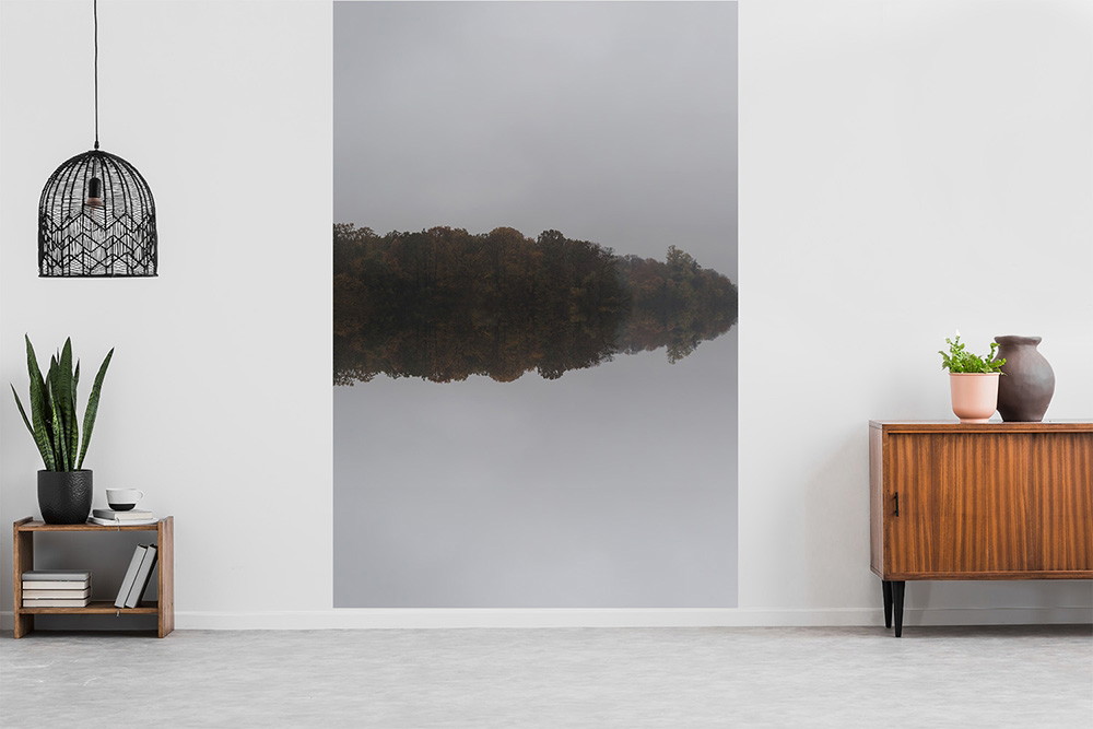 Wall mural - An atmospheric wallpaper shows a calm lake reflecting a misty forest on the surface of the water, embedded in a modern room design with plants and wooden furniture.