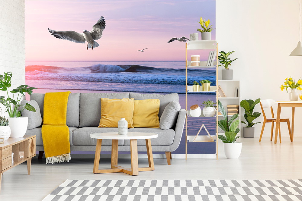Fototapete - Modernes Wohnzimmer mit grauem Sofa, gelber Decke, weißen Möbeln und Wandtapete mit Möwen über einem welligen Meer bei Sonnenuntergang