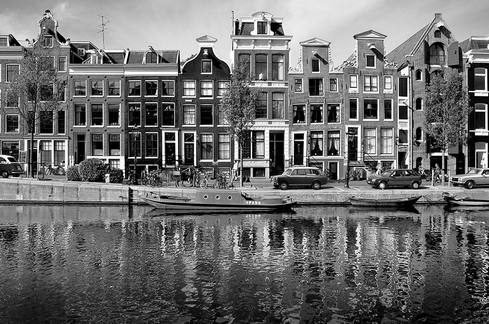 Papier peint photo - Photographie en noir et blanc de maisons historiques sur les canaux d'Amsterdam avec l'eau qui se reflète au premier plan
