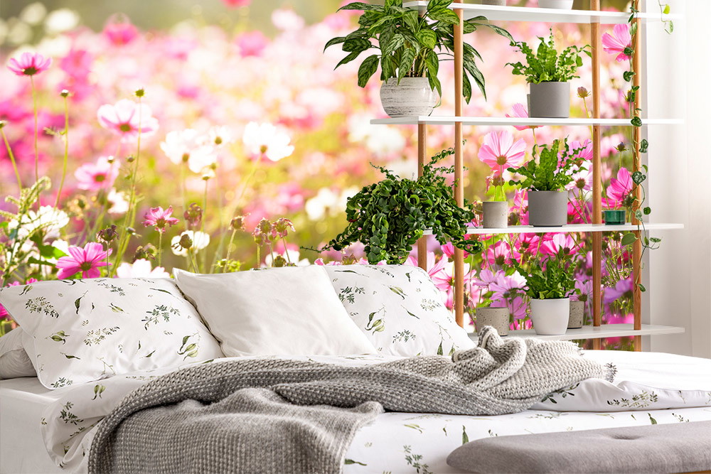 Fototapete - Schlafzimmer mit Blumenwiesenmotiv, weißen Kissen, grauer Decke und Pflanzen auf einem Regal