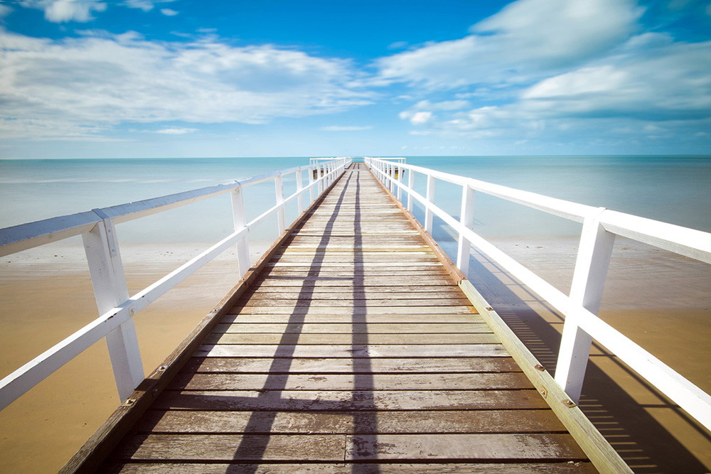 Fototapete - Ein langer Holzsteg erstreckt sich über einen sandigen Strand hinaus in das ruhige Meer unter einem klaren, blauen Himmel