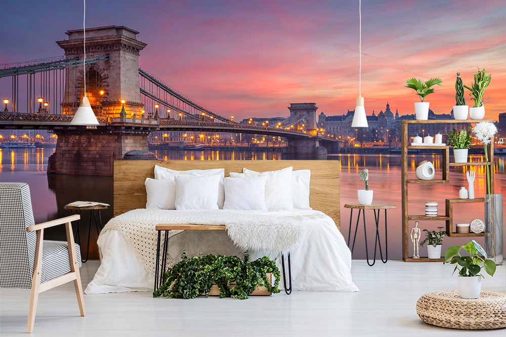 Fotobehang - Modern ingerichte slaapkamer met een groot bed, planten en op de achtergrond fotobehang van een verlichte brug bij zonsondergang