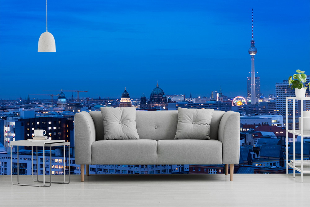 Papier peint photo - salon avec canapé gris devant l'impressionnante ligne d'horizon de Berlin la nuit, y compris la tour de télévision et les lumières brillantes.