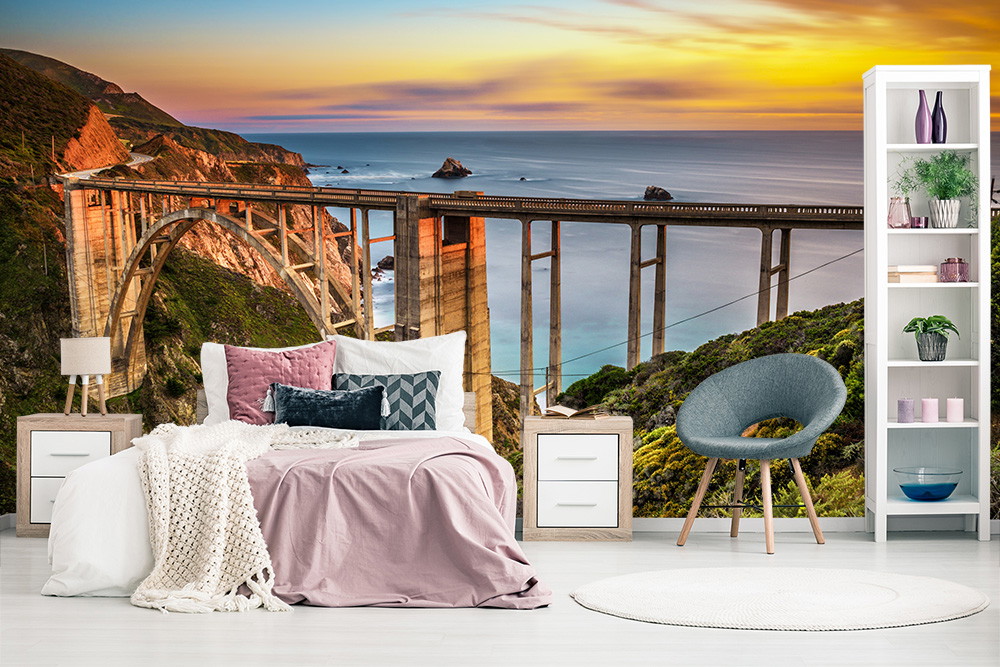 Fototapete - Schlafzimmer mit Bett und Sessel vor einer malerischen Küstenlandschaft mit Brücke im Sonnenuntergang