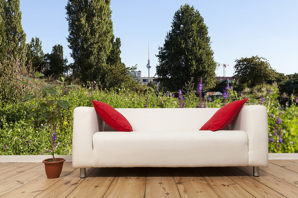 Fototapete - Helles Sofa mit roten Kissen vor einer Wand mit Gartenblick und Fernsehturm im Hintergrund