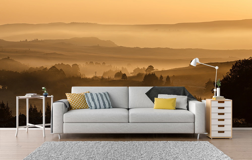 Papier peint photo - salon avec un papier peint représentant un coucher de soleil doré sur un paysage vallonné, avec un canapé blanc et des coussins décoratifs au premier plan