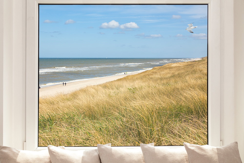 Fensterfolie - Aussicht auf einen Strand mit Dünen und Meer durch ein Fenster mit Kissen auf der Fensterbank