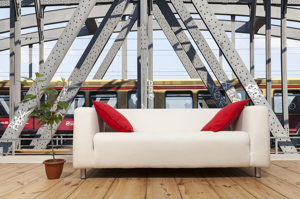 Fototapete - Weißes Sofa mit roten Kissen vor einer stählernen Brückenkonstruktion und vorbeifahrendem Zug