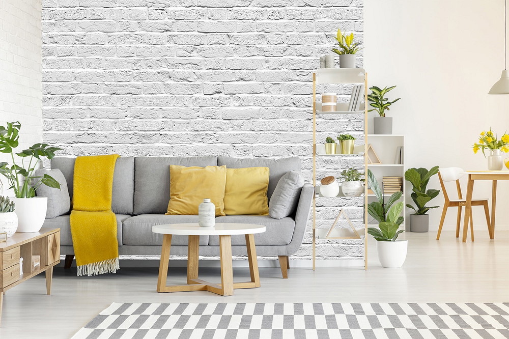 Fotomurale - Soggiorno elegante con divano grigio, cuscini gialli e coperta di fronte a una parete decorativa in mattoni bianchi, circondata da piante e scaffali