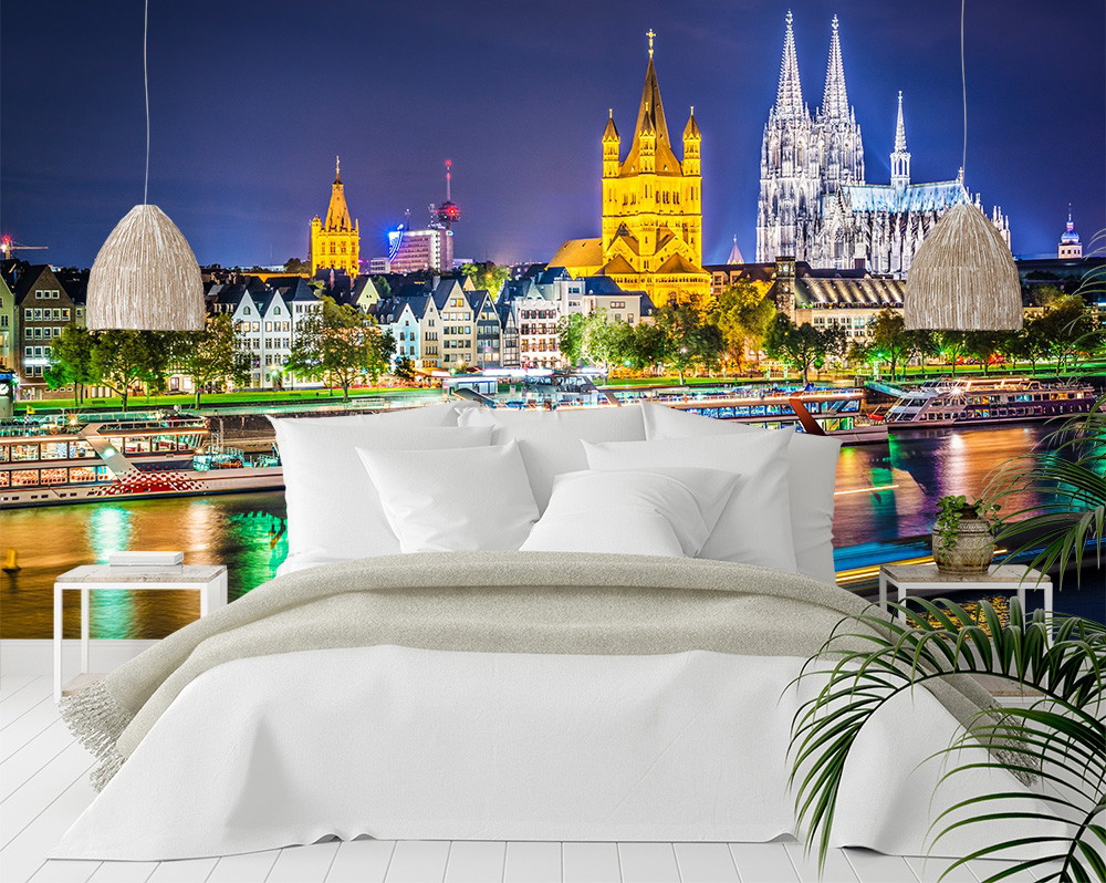 Papier peint photo - Chambre à coucher confortable devant une vue nocturne évocatrice de la skyline de Cologne avec la cathédrale illuminée et l'architecture historique au bord de la rivière