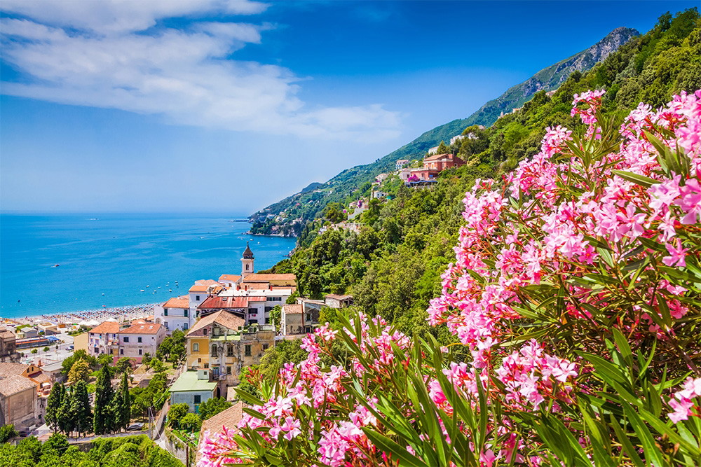 Fototapete - Blick auf eine idyllische Küstenstadt mit Oleanderblüten im Vordergrund, blauem Himmel und Meer