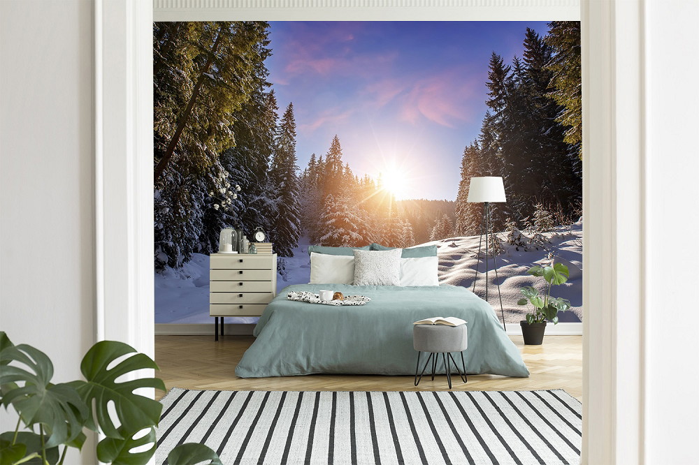 Fototapete - Schlafzimmer mit Bett und Nachttisch vor einer Fototapete, die eine verschneite Waldlandschaft mit Sonnenaufgang zeigt.