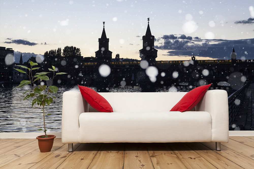 Wall mural - White couch with red cushions and potted plant in front of a city view with snowfall, in the background a bridge and evening sky