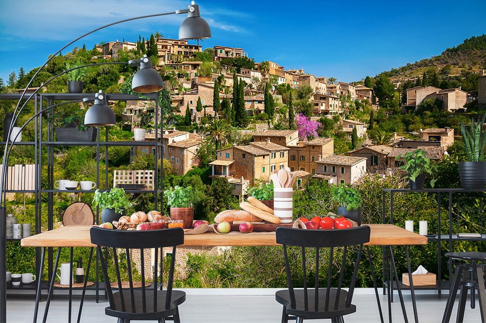 Muurschildering - Panoramisch uitzicht op een mediterraan dorp met stenen gebouwen en weelderige vegetatie op de achtergrond van een stijlvol ingerichte eetkamer met tafel, stoelen en decoratie.