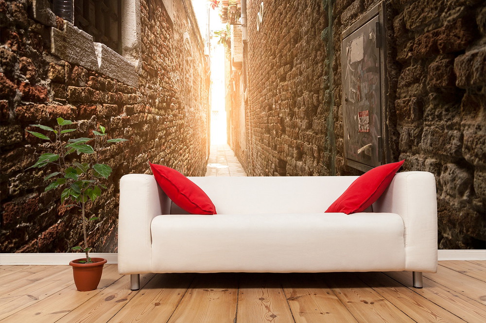 Fototapete - Weißes Sofa mit roten Kissen vor einer Tapete, die eine schmale, sonnenbeschienene Gasse aus Steinmauern zeigt, daneben eine Topfpflanze auf Holzboden
