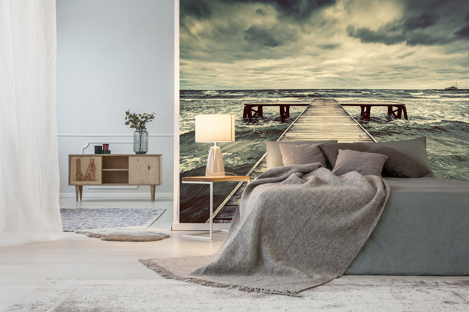 Fototapete - Schlafzimmer mit grauer Bettwäsche und Tapete eines Holzdocks im Meer unter wolkigem Himmel