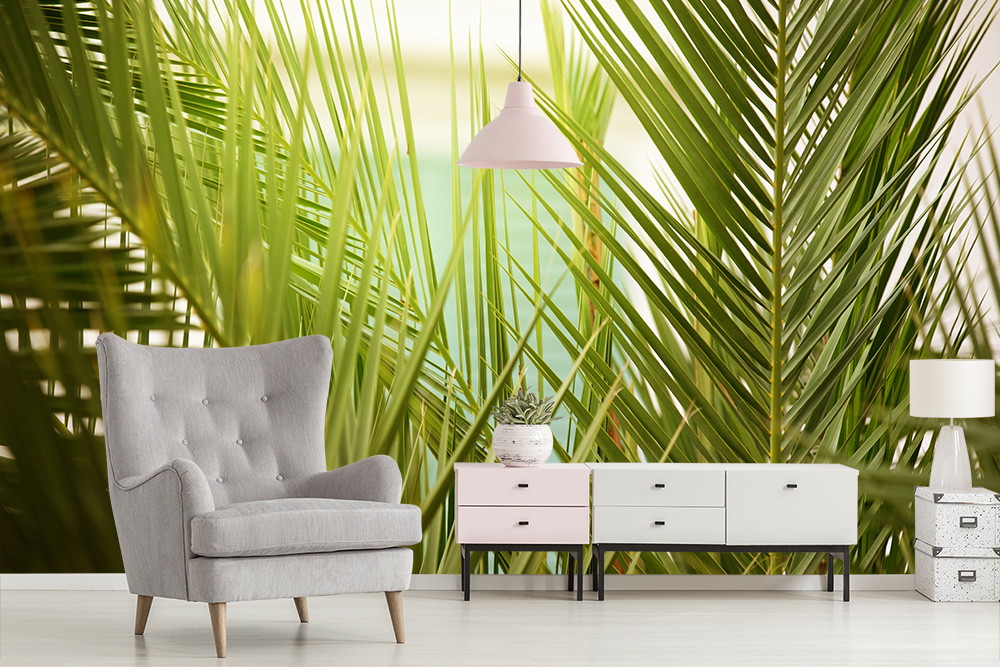 Fototapete - Wohnzimmer mit großer Fototapete, die ein dichtes, grünes Palmenblattmuster zeigt, eingerahmt von einem grauen Sessel und modernem Möbelstück.