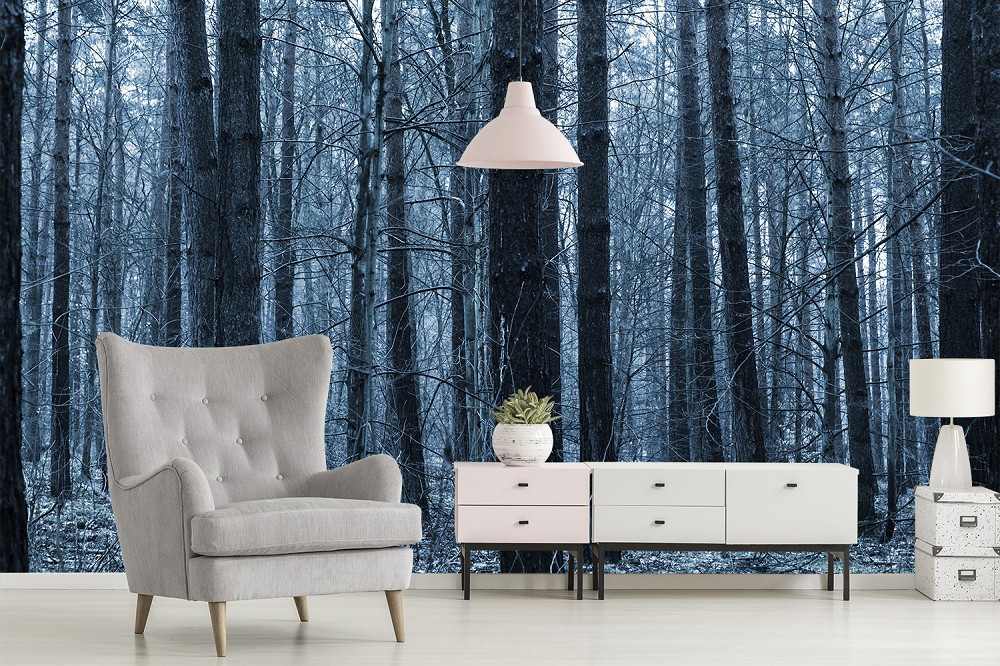 Fototapete - Waldmotiv in dunklen Blautönen hinter einem modernen Wohnzimmer mit grauem Sessel, Kommode, Lampe und Zimmerpflanze