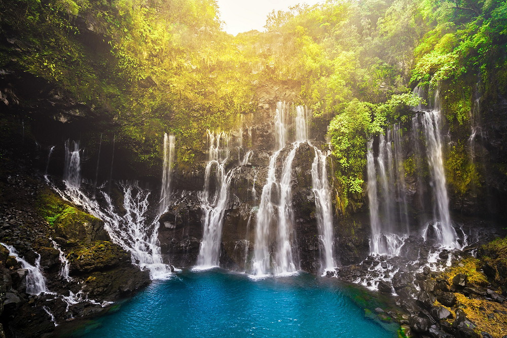 Fototapete - Ein majestätischer Wasserfall fließt in einen klaren, blauen Pool, umgeben von dichter, grüner Vegetation und hellem Sonnenlicht, das die Szenerie beleuchtet.