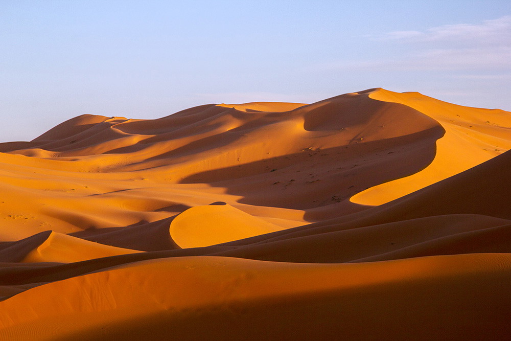 Fototapete - Wellenartige Sanddünen einer Wüstenlandschaft im warmen Licht der untergehenden Sonne