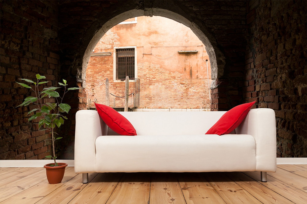 Fototapete - Wohnzimmer mit weißem Sofa, roten Kissen und Topfpflanze vor Ziegelwand mit venezianischem Fenster