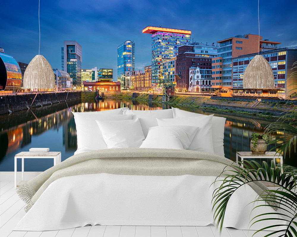 Fototapete - Beleuchtete Skyline einer modernen Stadt bei Nacht, die sich im Wasser spiegelt, hinter einem stilvollen weißen Bett platziert
