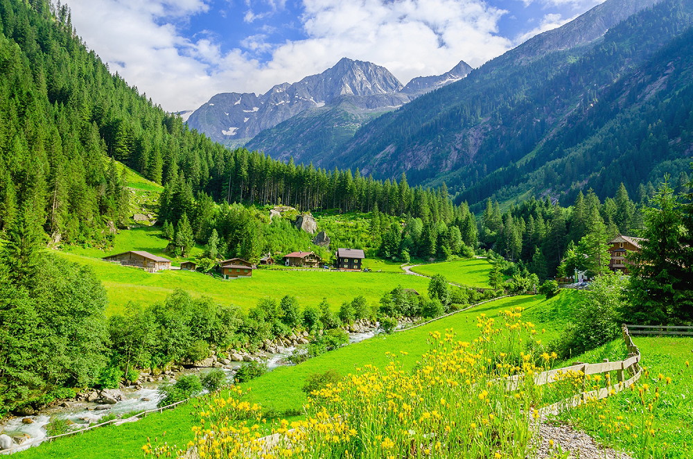Fototapete - Alpenidylle mit grünen Wiesen, Hütten und einem Fluss vor beeindruckenden Berggipfeln