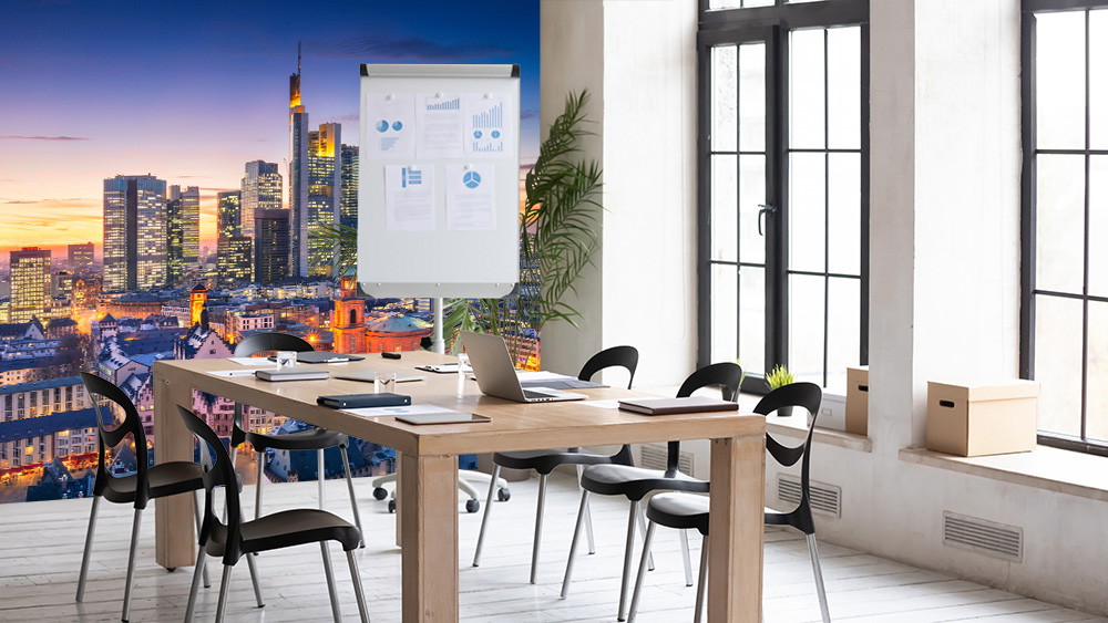 Fototapete - Moderner Besprechungsraum mit Tisch, Stühlen und Flipchart, Fensterfront mit Blick auf beleuchtete Skyline bei Sonnenuntergang im Hintergrund