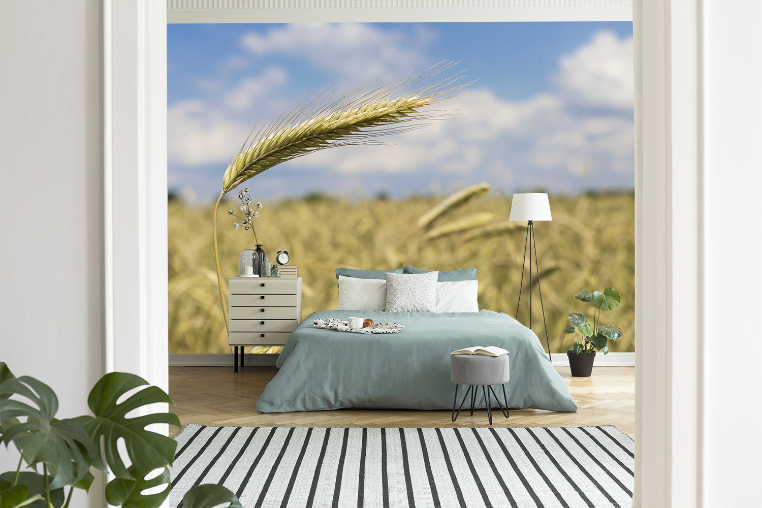 Fototapete - Ein Schlafzimmer mit einer Wandtapete, die ein Weizenfeld und blauen Himmel zeigt, ausgestattet mit einem Bett, Nachttisch und Pflanzen
