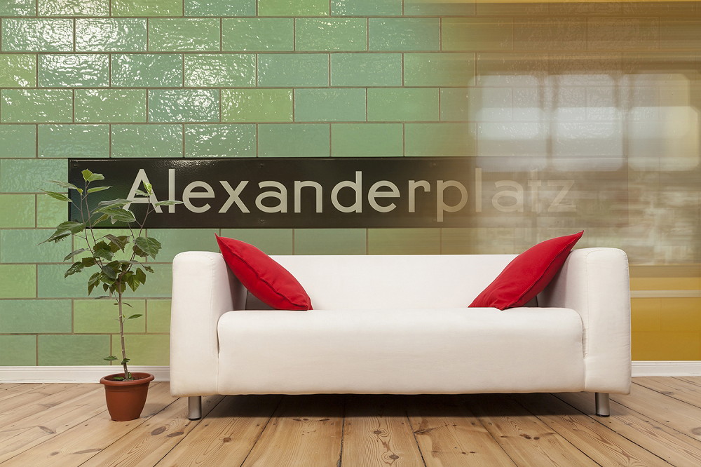 Fototapete - Weißes Sofa mit roten Kissen vor einer Wand mit Alexanderplatz-Schriftzug und Grünpflanze