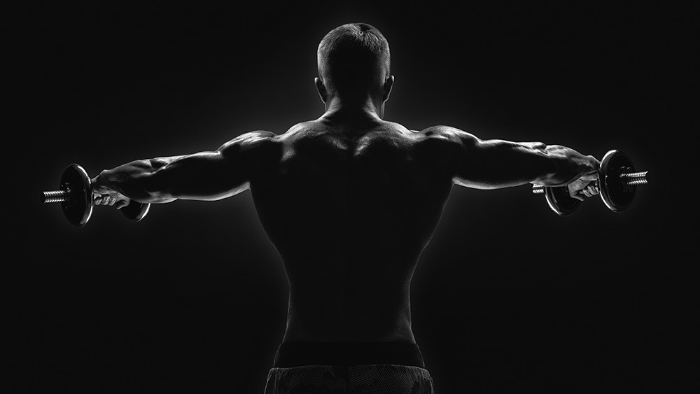 Wall mural - Back view of a muscular man lifting dumbbells in an atmospheric black and white shot