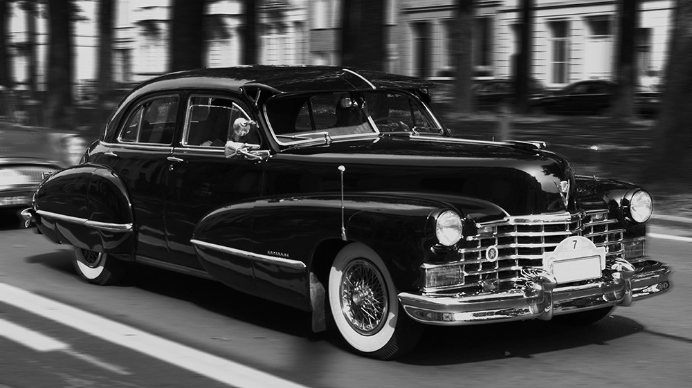 Fototapete - Schwarzer Oldtimer fährt dynamisch auf einer Allee, umgeben von Bäumen und anderen Fahrzeugen