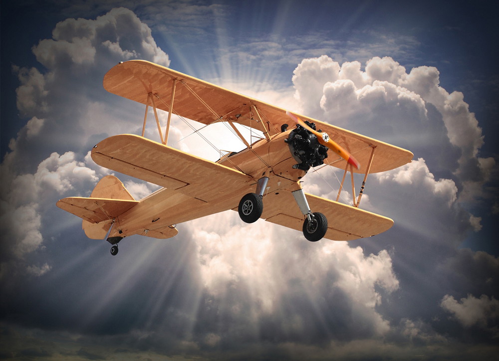 Fototapete - Ein vintage Doppeldeckerflugzeug fliegt vor einem dramatischen Himmel mit strahlenden Wolken