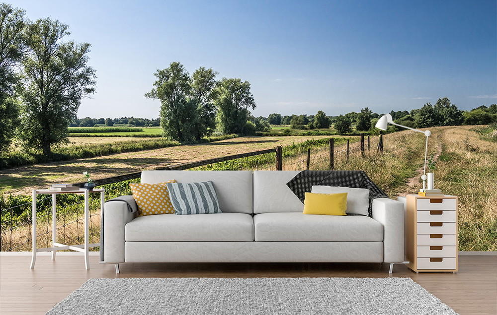 Muurschildering - Gezellige woonkamer met bank, ervoor een landschap met weilanden en bomen