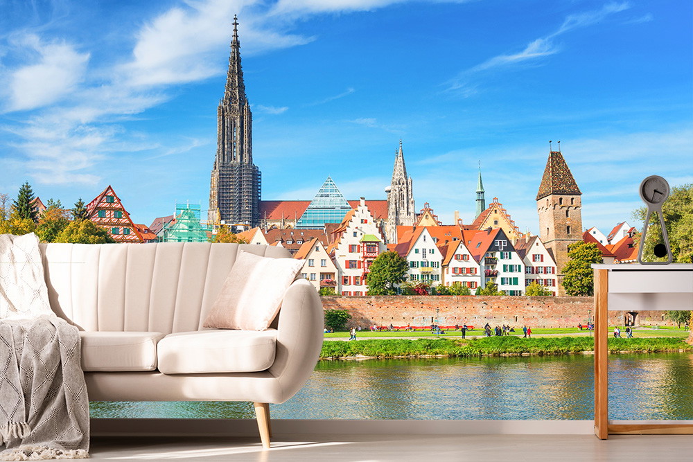 Fotomurale - Veduta di Ulm con la cattedrale di Ulm e il Danubio in primo piano, incorniciata da una scena di soggiorno moderno con divano e tavolo