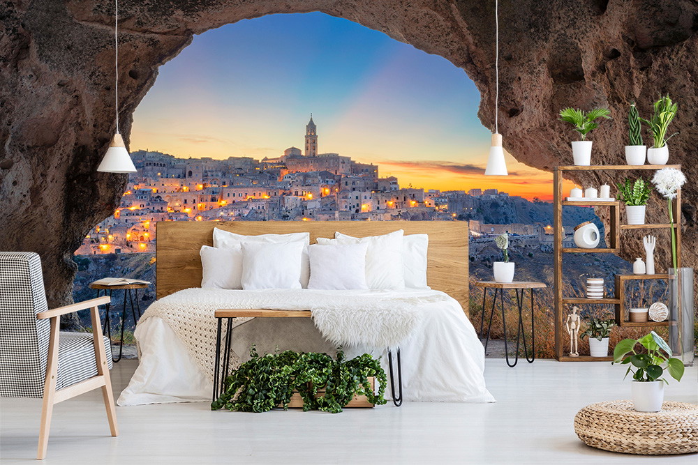 Wall mural - Bedroom with a view of a picturesque cityscape at sunset, framed by a cave opening, decorated with plants and modern furniture