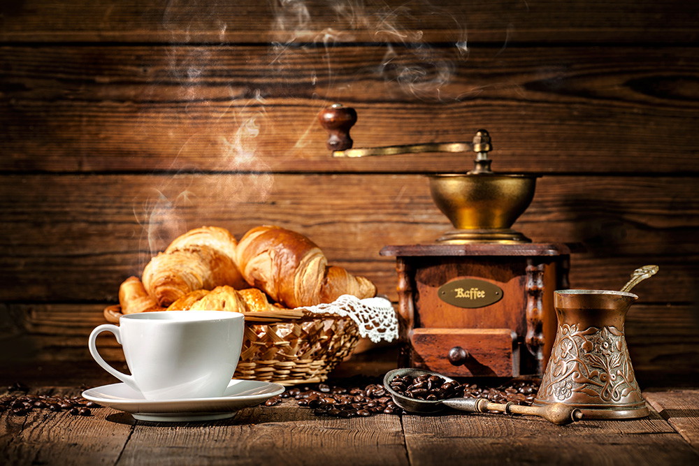 Muurschildering - Een rustiek ontbijtarrangement met dampende koffie, verse croissants, een koffiemolen en koffiebonen.