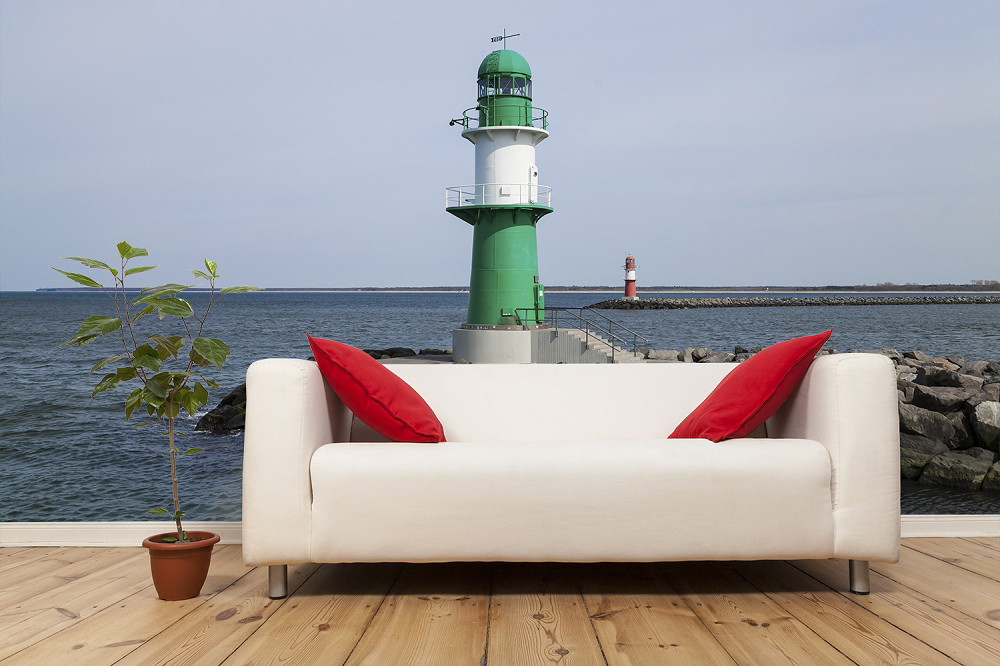 Fotobehang - Woonkamer met bank, rode kussens, potplant en uitzicht op een vuurtoren aan zee
