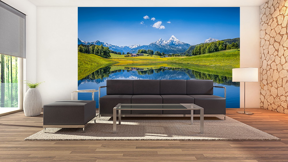 Fotomurale - Arredamento moderno per il soggiorno con grandi finestre e una carta da parati di grande effetto che raffigura un paesaggio montano verde con un cielo azzurro e un lago tranquillo.