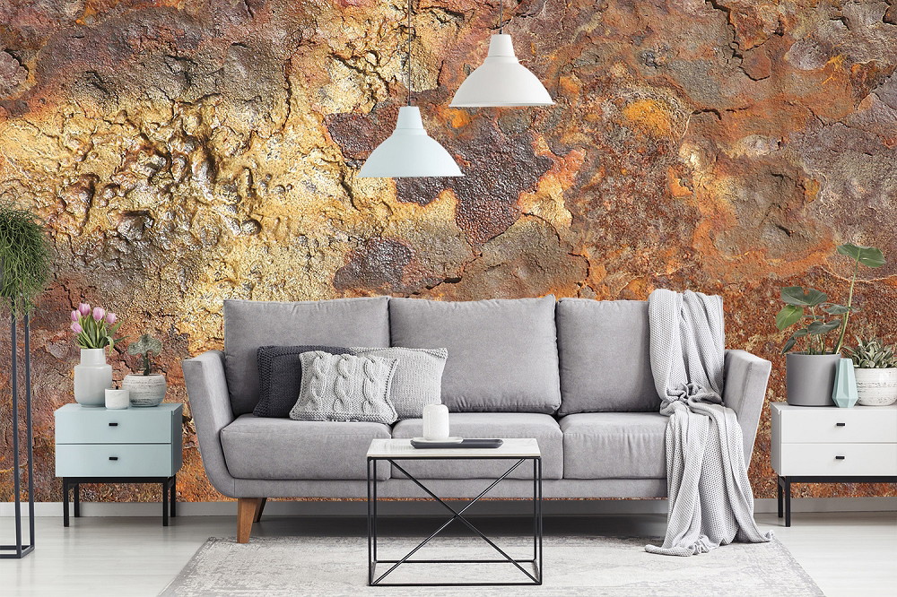 Fototapete - Ein modernes Wohnzimmer mit einem grauen Sofa, umgeben von Pflanzen und Lampen, vor einer Wand in rustikaler Steinoptik.