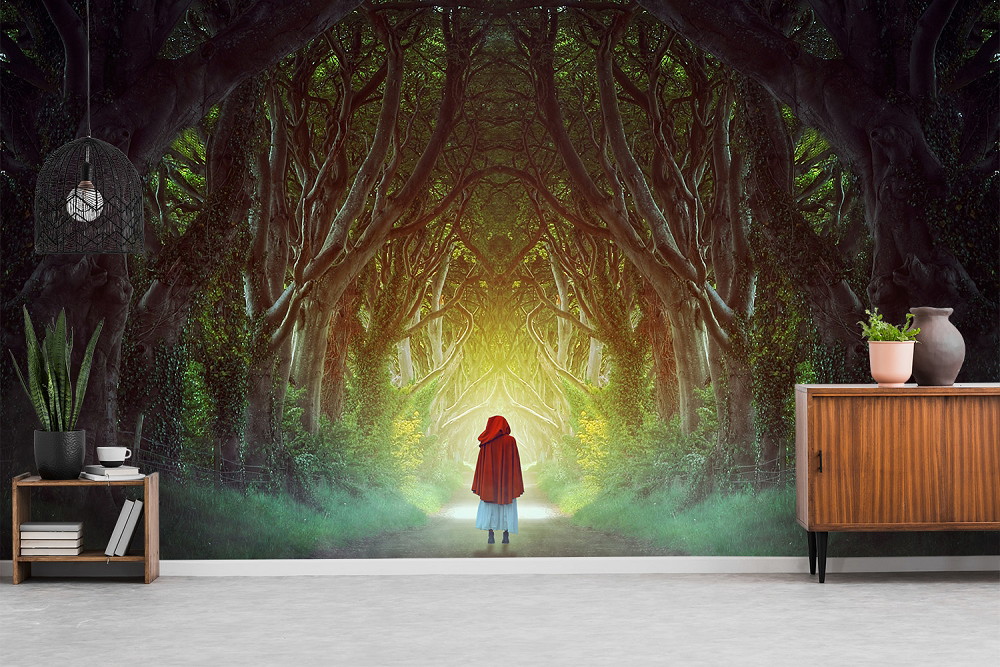 Fotomurale - Un bambino con un mantello rosso si trova su un sentiero costeggiato da alberi alti e contorti, che creano un'atmosfera misteriosa di foresta