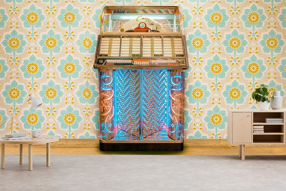 Fototapete - Eine stilvolle Vintage-Jukebox steht vor einer Wand mit retro-blumiger Tapete. Daneben befinden sich ein Holztisch mit Lampe und eine Kommode, die einen gemütlichen Raum aus den 70er Jahren nachbilden.
