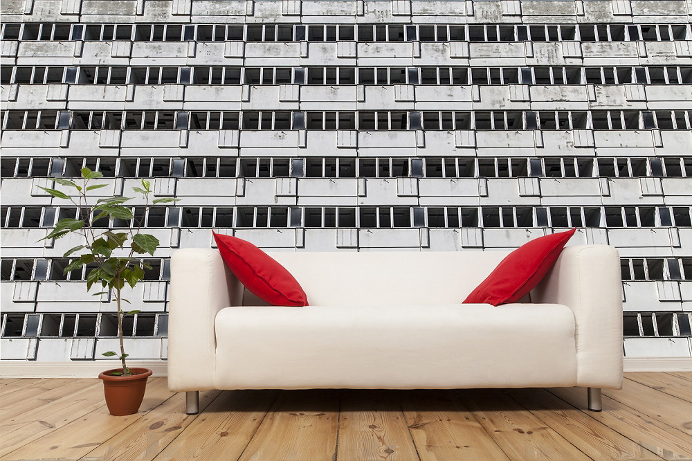 Fototapeta - Biała sofa z czerwonymi poduszkami przed tapetą z motywem miejskiej fasady, obok rośliny doniczkowej