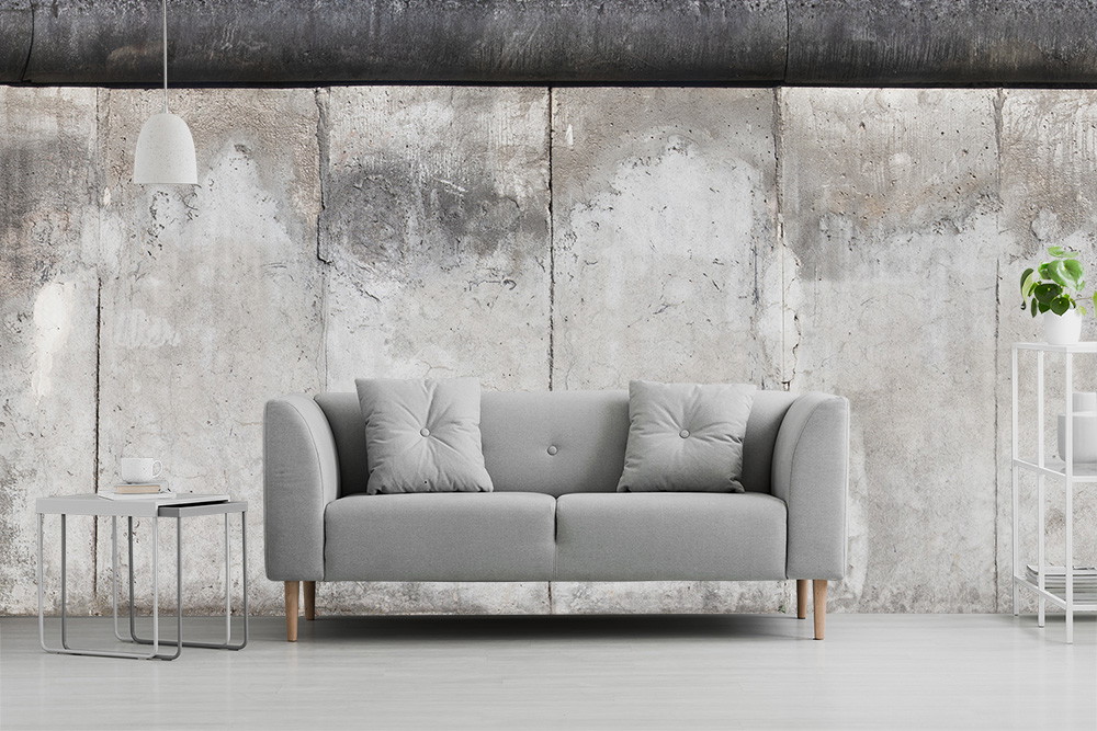 Fototapeta - Szara sofa przed betonową ścianą, otoczona bocznym stolikiem i półką z rośliną.